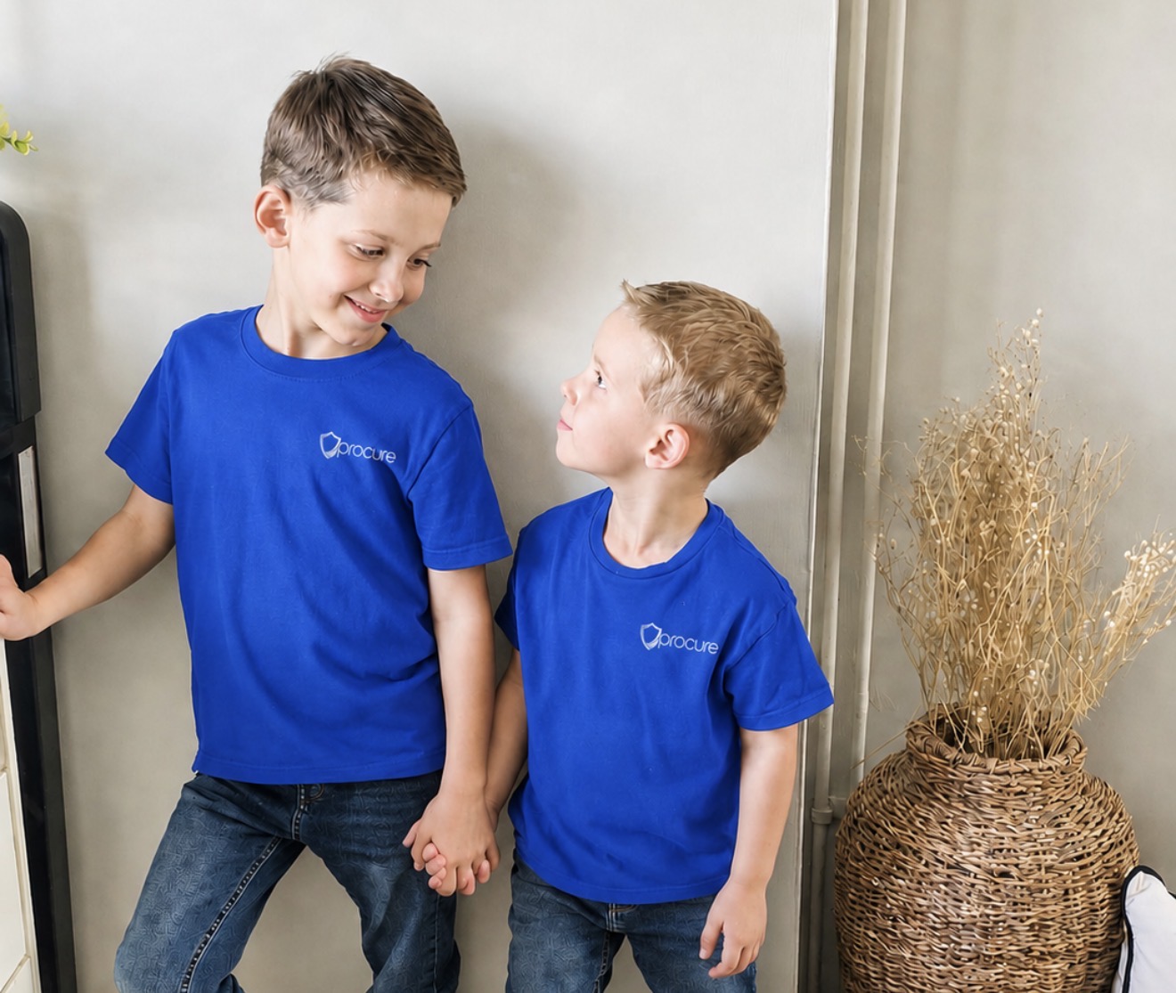 Two young children wearing Procure Group branded t-shirts, holding hands at home